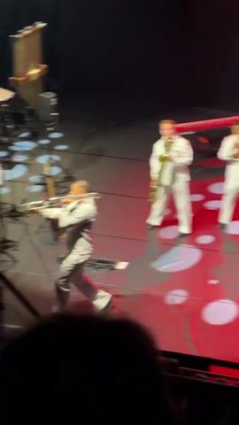 The video shows a live musical performance on a stage. In the foreground, a person wearing a white uniform and a grey vest is playing a trombone. They are positioned in the center of the stage and appear to be actively performing, leaning into the instrument. To the right of the trombonist, a person in a white uniform is playing a saxophone. Behind the trombonist, to the left, several other musicians are seated, also in white uniforms. One is playing a large brass instrument, possibly a tuba, and another appears to be seated at a drum set. Various musical equipment, including microphones, stands, cables, and speakers, are visible on the stage. The stage floor has a red design with white circular patterns. Lighting from above illuminates the performers. In the lower part of the frame, the backs of audience members can be seen. The overall environment suggests a concert hall or theater.