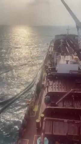 The video shows the side of a large red ship in the ocean on a hazy day. Multiple long, dark hoses are partially submerged in the water next to the ship. The sun reflects on the water's surface. The camera pans slightly to follow the movement of the hoses in the water.