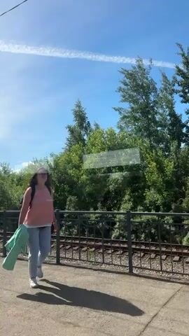 A woman in a pink shirt and gray pants walks along a train platform, carrying a green jacket and a backpack. She is looking to her right. Behind her, train tracks run parallel to the platform, bordered by a black metal fence. Beyond the tracks, lush green trees and foliage are visible under a bright blue sky with a white contrail. A man wearing a tan shirt, shorts, a baseball cap, and a mask walks towards the camera. He is carrying a bag in his left hand. The sun is shining, casting shadows on the platform.