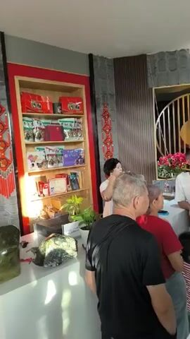 A group of people are gathered in a room with a decorative display shelf on the left. The shelf is red and contains various items, including boxes, bags, and books. Two red and gold decorative ornaments hang on the wall to the left of the shelf. In the center of the room, a woman with short black hair is speaking to a man in a white t-shirt. Several other individuals are also present, some facing the speakers and others looking away. The environment appears to be indoors, possibly a shop or a gathering space, with textured walls and a wooden paneling on the right.