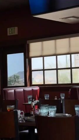 The video shows the interior of a restaurant. In the foreground, there are tables set with chairs and glasses. In the background, there is a window with blinds, through which a desert landscape is visible. To the left of the window, there is a door with an "EXIT" sign above it. Sunlight streams into the restaurant, casting shadows on the tables.