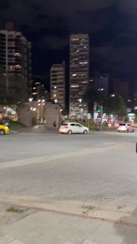 The video shows a cityscape at night. Buildings rise in the background, lit by artificial lights. A street with brick paving occupies the foreground. A yellow taxi and a white car drive on the street. A grassy park area is adjacent to the street, where many people are sitting and standing, appearing relaxed. Trees are interspersed throughout the park. Pedestrians are also visible on the sidewalks and at the edges of the park. The overall atmosphere is calm and social.