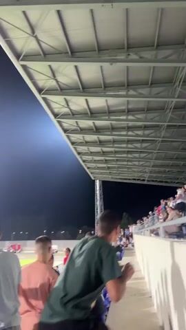 The video shows a view from the stands of a sports stadium at night. The camera pans slightly to the left. In the foreground, several people are visible, some standing and some sitting. They appear to be spectators at an event. The stadium lights are on, illuminating a bright green field in the background. The sky above is dark. The overall atmosphere appears to be one of a sporting event or concert.