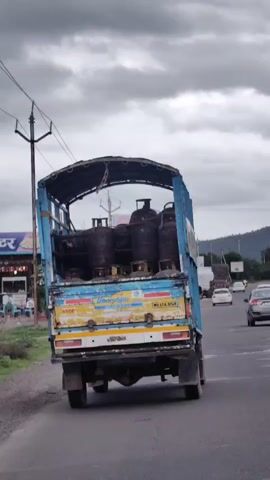 The video shows the rear of a blue and yellow truck loaded with gas cylinders. The truck is on a road, with other vehicles visible in the background. The truck has "Bharatgas" written on the back. The sky is cloudy.