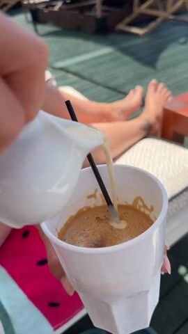The video captures a serene, sunny day at a beachside location in Νέα Χηλή, Greece.

The initial frames show a close-up of a person's hands pouring creamer from a small white ceramic pitcher into a white faceted cup containing coffee and a black straw. In the background, another person's bare legs and feet are visible, resting on a lounge chair on a green wooden deck, suggesting a relaxed atmosphere. A red and white striped towel or fabric is partially visible under the chair.

As the video progresses, the view widens to reveal the beautiful beachfront setting. Two large, stylish lounge beds with natural-colored fringed canopies are prominently featured on the wooden deck, offering shade. A small wooden side table between them holds a white bucket, a wooden ashtray, and some papers. Beyond the deck, the calm blue sea stretches out under a clear blue sky, meeting a sandy beach. In the distance, an adult and a child can be seen walking along the water's edge.

The overall scene depicts a peaceful and leisurely moment, likely at a beach club or resort, where individuals are enjoying coffee while relaxing by the sea on a bright, beautiful day.