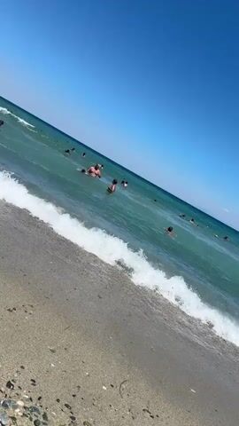 A handheld, slightly angled video captures a sunny day at a beach. The foreground shows a sandy shore with some small pebbles and debris. A gentle wave is breaking and rolling onto the beach. The ocean water is a clear turquoise color. In the middle ground and background, numerous people are swimming and enjoying the water. They are spread out across the visible area of the sea. The sky is a clear, vibrant blue, indicating fair weather. The overall impression is one of a peaceful and lively day at the beach.