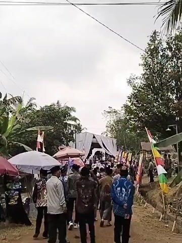 A group of people are gathered outdoors in what appears to be a village or rural setting. They are walking along a dirt path towards a white tent structure which is decorated with flags and banners. Some people are wearing traditional clothing, including head coverings. There are trees and vegetation surrounding the area. The sky is overcast. There are several market stalls with umbrellas set up on the side of the path, with goods displayed for sale.