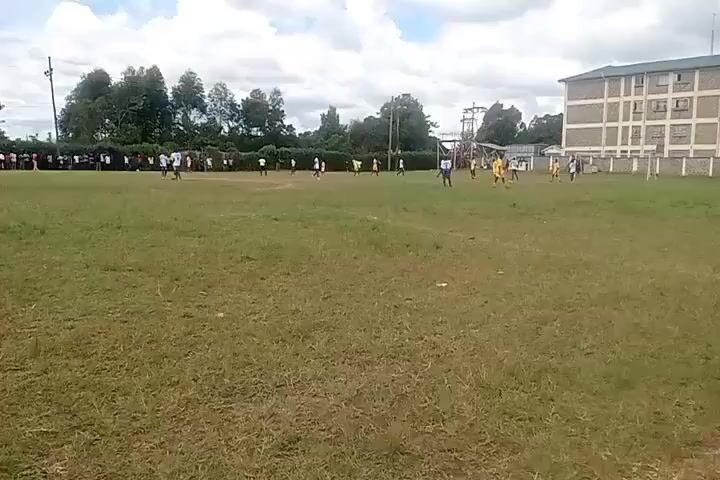 A wide shot captures a soccer match underway on a grass field under a partly cloudy sky. There are two teams, one in white and the other in yellow.  The white team is closer to the viewer, lining the sidelines. There's a building in the background on the right, and trees in the background to the left.