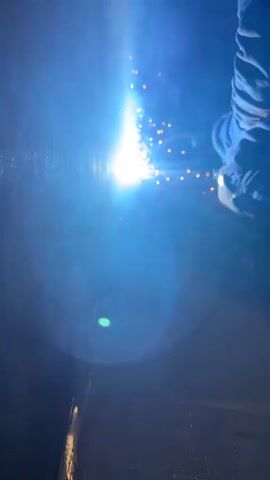 A welder wearing a helmet and protective clothing is working on a metal surface. The bright light of the welding arc illuminates the scene, casting sparks and smoke. The welder appears to be in the process of joining two pieces of metal. The environment is dark, suggesting an indoor or nighttime setting.