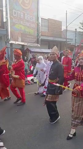 A group of people in traditional attire are walking down a street in a procession. The people are dressed in colorful clothing, with some wearing head coverings and others wearing hats. Some of the individuals are carrying flags. In the background, there are buildings and a large banner with text in a foreign language. The overall atmosphere appears festive.
