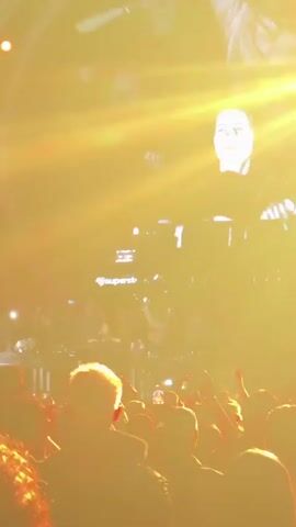 The video shows a concert crowd with hands raised. A stage is visible in the background with lights and a large circular screen displaying abstract visuals. The lighting is bright and predominantly yellow and orange, creating a warm and energetic atmosphere. Some people are holding up their phones, presumably to record or take pictures. The scene appears to be dynamic and lively, characteristic of a live music performance.