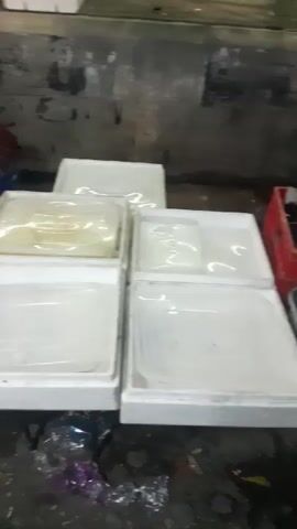 The video shows a man in a black t-shirt and black gloves sitting on the ground and cutting a melon. He is surrounded by several white styrofoam containers, some of which are filled with sliced melon. There are also whole melons on a cutting board. The background appears to be a market stall with concrete walls and a metal counter. The ground is wet and appears to be dirty.