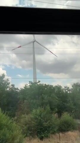 The video shows a view through a window of a wind turbine and surrounding vegetation. The wind turbine is tall and white with three blades. The blades are long and thin, with red and white markings on the tips. The turbine is set against a backdrop of a cloudy sky with patches of blue. In the foreground, there is lush green vegetation, including trees and bushes. There are also some power lines visible in the frame. The camera appears to be moving, as the view shifts slightly between frames.