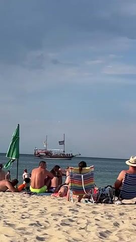 The video shows a busy beach scene in Kiten, Bulgaria, on a sunny day with some clouds in the sky. In the foreground, several people are relaxing on the sand, some sitting on towels and others in beach chairs. One person is wearing a hat, and another is in a colorful striped beach chair. A green beach umbrella is also visible.

In the middle ground, a large boat with a pirate flag is sailing on the turquoise water. The boat appears to be carrying many passengers, suggesting a tour or excursion. The water is calm and clear, reflecting the sky.

The overall mood is relaxed and summery, with people enjoying a day at the beach. The time of day appears to be afternoon, given the lighting and the long shadows. The location is clearly identified as Kiten, Bulgaria. There is no visible text in the video.