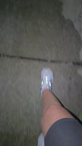 The video shows a first-person perspective of someone walking on a wet, textured surface, possibly a sidewalk or pavement. The individual is wearing athletic shoes, one of which is clearly visible with a reflective detail on the heel. Dark shorts are also visible on the lower half of the frame. The ground is wet and appears to have dark, uneven patches, possibly due to water or a stain. The camera is moving, suggesting the person is in motion. The lighting appears dim, indicating it might be nighttime or a poorly lit area.
