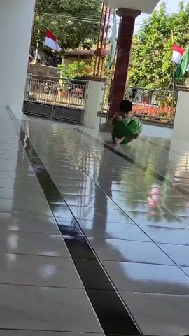 A young boy wearing a black cap and green pants and a light-colored shirt sits cross-legged on a tiled floor, looking down at something in his hands. The floor is polished and reflects the surroundings. Behind him, a fence separates the tiled area from a garden with trees and plants. Two flags, one red and white and another green and white, are visible on poles. The overall impression is of a sunny day, possibly outdoors or in a covered porch area.