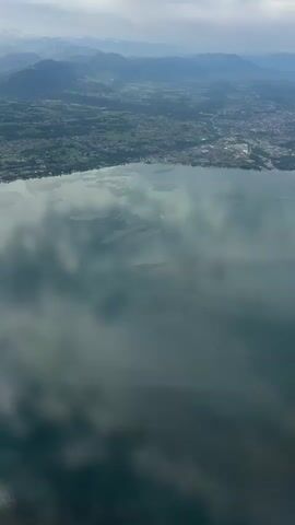 The video shows an aerial view of a large body of water, possibly a lake, with a town or city along its edge. Mountains are visible in the background. The water's surface reflects the sky, creating a mirrored effect. The overall scene is captured from a high vantage point.