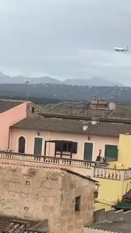 The video shows a commercial airplane in the sky, on approach to land. The airplane is white with blue accents, and it is flying against a grey, overcast sky. Below the plane are rooftops of buildings, primarily pale shades of pink and yellow, with tile roofs. In the distance are mountains, also shrouded in grey. The setting appears to be a residential area with older architecture.