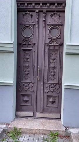 The video begins with a close-up of a decorative, double wooden door. The door is dark brown with ornate carvings, including circular insets with numbers 13 and 96, and diamond shapes. The door has a brass handle and a metal plate at the bottom. To the left of the door is a light green wall with a gray base and a window. The camera then pans to the right, showing more of the building's exterior. The building has light green walls with white trim, and multiple windows. Some windows are open. There are balconies on upper floors with black railings. The ground in front of the building is covered with grass and some small plants. The video ends showing a wider view of the building, revealing its architectural style, which appears to be old European.