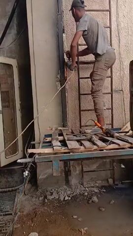 A man stands on a platform constructed of wooden planks and a blue metal frame, using an angle grinder to cut into a metal wall. The platform is positioned in front of an electrical cabinet with an open door. The man is wearing a gray t-shirt, cargo pants, and a dark baseball cap. Sparks fly from the grinder as he cuts. He is also standing on a small ladder. The ground beneath the platform is cluttered with debris.