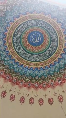 The video shows an upward view of an ornate ceiling with Islamic calligraphy and geometric patterns in concentric circles. The innermost circle contains the word "Allah" in Arabic script, centered in a blue disc. Surrounding this are several rings of calligraphic text and decorative patterns in shades of blue, green, red, and gold. The outermost area of the ceiling features a repeating pattern of teardrop-shaped motifs. The video is taken from below, suggesting the viewer is looking up at the decorated ceiling. The lighting is bright, possibly from natural light entering the space. An audio track accompanies the visual.