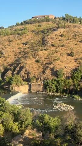 A wide shot captures a river flowing through a valley, bordered by lush green trees and a dry, scrub-covered hillside. A dilapidated stone structure, possibly a dam or remnants of an old building, is visible on the left bank of the river. The hillside is dotted with patches of dry grass and scattered green vegetation. Several trees stand on the right bank of the river, with dense foliage. In the distance, atop the hill, several buildings and structures are visible, suggesting a settlement or town. The sky is clear and blue, indicating fair weather and daylight. The video appears to be a panning shot, moving from left to right, offering a panoramic view of the landscape. There are no visible people in the frame.