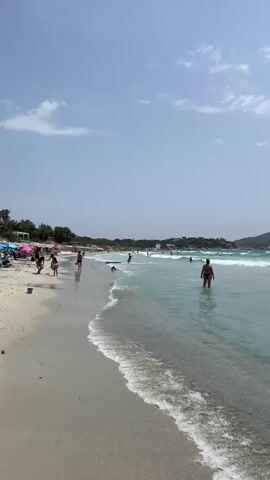 The video shows a beach scene with people swimming and walking in the shallow water. The water is clear and turquoise, with gentle waves lapping at the sandy shore. In the background, there are sailboats and a distant hillside. The sky is blue with a few scattered clouds. The overall atmosphere is one of relaxation and summer fun.