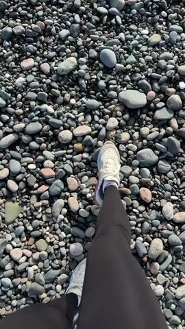 The video shows a person walking on a pebbled beach, with the camera angle from above, focused on their lower legs and feet. The person is wearing black leggings and white sneakers. The beach is covered with an array of small, smooth stones in various shades of grey, brown, and pink. As the person walks, the camera pans upwards to reveal waves breaking on the shore, creating white foam. The sea stretches to the horizon under a sky with a few scattered clouds. The scene suggests a walk along the beach on a day with gentle waves.