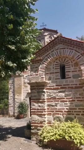 The video shows an exterior view of an old stone and brick church on a sunny day. Lush green trees and bushes surround the building, casting shadows on the ground. The architecture features arched windows and intricate brickwork patterns. A cross is visible on the dome of the church. The overall impression is one of tranquility and historical significance.