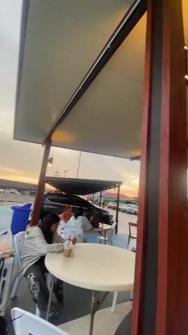 The camera pans across a covered outdoor seating area.  Numerous people are sitting at round white tables, some eating and drinking, others looking at their phones or talking. There are suitcases and backpacks near some of the tables.  Outside the seating area, cars are parked and the sky is bright. The seating area has a low white ceiling with dark trim. There are dark wooden posts throughout the seating area.