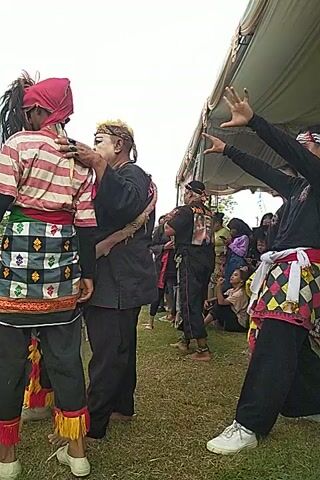 Here is a concise description of the video:

A group of people wearing traditional clothing are performing a dance or ritual. They are standing on a grassy field, with a tent structure visible in the background. Some members of the group are in close contact, embracing each other. Other individuals, who appear to be spectators, are watching in the background. The video is shot from a low angle and seems to be rotated or filmed sideways.