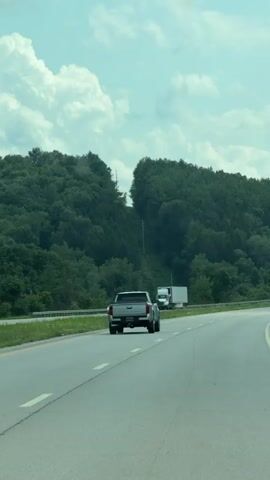 A silver pickup truck travels on a highway, followed by a white box truck in the distance. The highway curves to the right, bordered by a grassy shoulder and a dense forest. Two large white cumulus clouds are visible in a clear blue sky above the tree-covered hills.