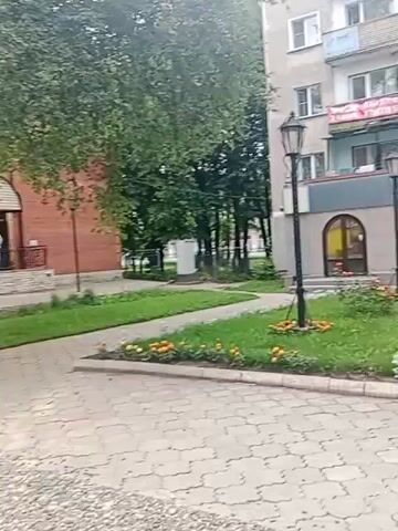 The video shows a street view with a building and a path. The building has a sign that says "La DONNA" above arched windows. There are two street lamps in front of the building. The path is paved with interlocking bricks. Green grass and some flowers are visible on the left side of the path. Trees with green leaves are on the right side of the path, and part of a brick building is visible behind the trees. Further down the path, there are people walking and sitting. The sky is overcast.