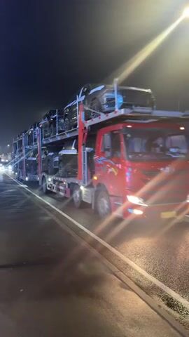 The video shows a car carrier truck driving on a road at night. The truck is carrying multiple cars on a multi-level trailer. The cars are dark in color and appear to be new. The truck has a red cab with a flame design on the side. Streetlights illuminate the road. The camera is stationary and captures the truck as it passes by. The overall environment is dark due to the nighttime setting.