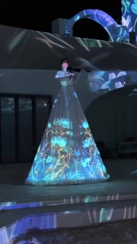 A woman in a long white dress stands on a slightly elevated platform at night, playing a violin. The dress is illuminated with projected blue and yellow flower-like designs. Similar floral projections are also cast onto the building behind her, including an arched doorway. The platform she stands on is part of a multi-tiered staircase with similar floral projections on the steps. The environment is dark except for the projections and some ambient lighting on the building.