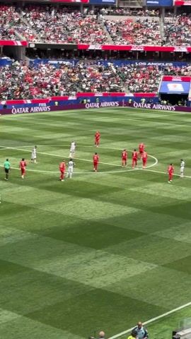 A high-angle, medium shot shows a soccer match in progress. In the foreground, players in red and white uniforms are scattered across a bright green field, some standing still and others walking. One player in a red uniform with the number 9 on his back walks away from the camera, followed by a referee in a green shirt and black shorts. The field is marked with white lines and has the "QATAR AIRWAYS" logo visible along the sidelines. In the background, a stadium filled with spectators wearing red and white clothing is visible. The stands are tiered and brightly lit. A large banner displaying "FIFA WORLD CUP 2026" is also visible in the background. The overall scene suggests a professional soccer game taking place in a large, outdoor stadium.