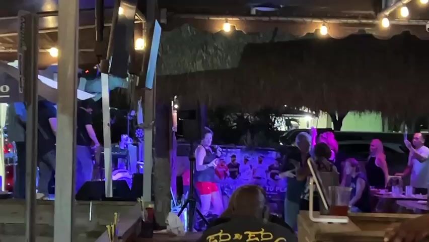 A band performs on a stage with blue and purple lights, while a singer in a red skirt stands at a microphone. People watch and dance in front of the stage. The scene appears to be an outdoor venue with a thatched roof and string lights.