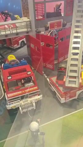 The video shows a close-up view of a detailed Playmobil firefighter station playset. In the foreground, a red fire truck with flashing blue lights is parked. A small firefighter figurine is attached to a hose extending from the truck, seemingly in the process of spraying water. To the right of this truck, another red fire truck with an extendable ladder is parked near the station building. The building itself is multi-story and red, with large garage doors. A tall, silver ladder extends from the side of the building. Several other Playmobil figurines are visible within the station and on the upper levels. The scene appears to be set on a green and grey surface representing a street or courtyard. The overall impression is of an active emergency response scenario within the toy set.