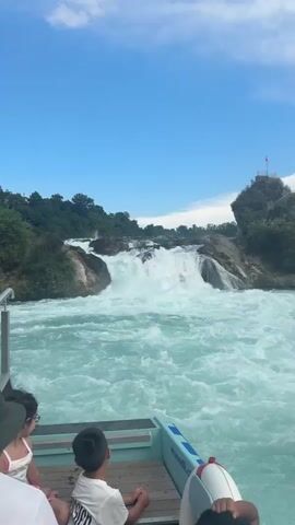 The video shows a boat moving towards a waterfall. People are seated on the boat, facing away from the camera, looking towards the waterfall. The water is turquoise and choppy, with white foam from the waterfall. The waterfall is wide and powerful, with water cascading over rocks. There are trees and greenery on the banks of the river. In the background, there is a large building on top of a hill overlooking the waterfall. A smaller yellow boat is visible near the waterfall. The sky is blue with some white clouds.