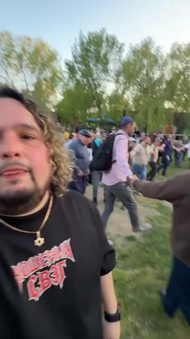 The video shows a man filming himself in a grassy field with a large crowd of people in the background, a bonfire, and an inflatable slide. The man in the foreground wears a black t-shirt, a gold chain, and has curly hair and a beard. Behind him, people are dancing in a circle around a bonfire. Some individuals are wearing traditional Jewish attire. An inflatable slide stands in the background. The scene appears festive and lively.