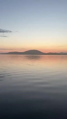 The video shows a body of water with ripples and reflections of a pale sky with pink and orange hues, indicating either sunrise or sunset. Hills or mountains are visible in the distance, silhouetted against the sky. On the right side of the frame, there is a shoreline with gravel and trees. The water appears calm and clear, with the shoreline also visible beneath the surface in some areas. The scene is peaceful and natural.