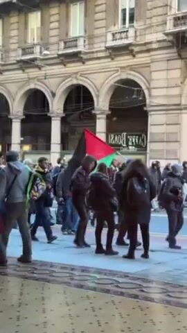 The video shows a crowd of people standing on a sidewalk in front of a building with arches. The people are of various ages and ethnicities. Many are wearing dark-colored clothing. A Palestinian flag is being carried in the crowd. Some people appear to be taking pictures or videos. The building behind them has a sign that reads "CAFFE BOASI." The sidewalk has a mosaic pattern.