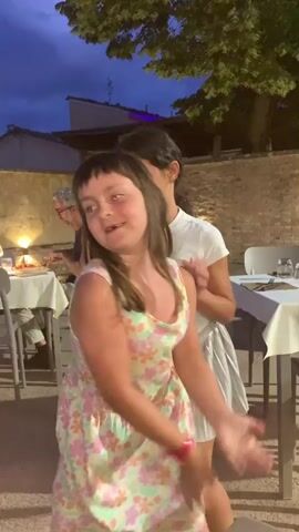 The video captures two young girls dancing outdoors at dusk. The girl in the foreground wears a floral dress with pastel colors and has her hair in pigtails. She is enthusiastically dancing, with her arms moving dynamically. Behind her, another girl in a white t-shirt and a silver pleated skirt is also dancing and smiling, her hands clasped in front of her chest. The setting appears to be an outdoor dining area of a restaurant. In the background, tables are set with white tablecloths and place settings. A man with gray hair is seated at a table, looking towards the left. A brick wall and a large tree with green foliage are visible behind the girls. The sky is a deep blue, indicating it is late evening or early twilight. The lighting suggests artificial lamps are present, illuminating the tables. The overall mood is joyous and playful, with the children clearly enjoying themselves. There is no visible text in the video.