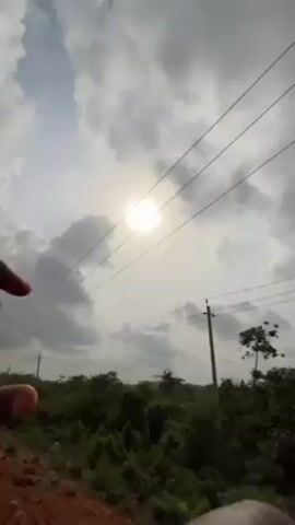 A person is holding their hand up to the sun, attempting to obscure it. The sun is partially obscured by clouds. The scene takes place outside, with a foreground of green vegetation and a background of utility poles and lines. The person is wearing a dark-colored shirt.