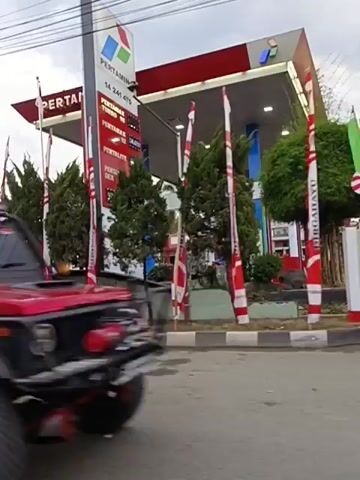 A black SUV drives past a Pertamina gas station. The gas station has a red roof and is decorated with flags. There are several cars and motorcycles in the vicinity. The scene is a street with buildings and overhead wires.