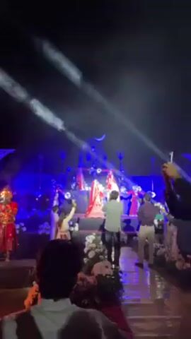 The video captures a lively event, likely a wedding celebration.  A bride dressed in red stands on a stage, illuminated by blue and white lights and surrounded by dancers in red attire. An image of Shiva, complete with a crescent moon, is projected in the background. Numerous people in the audience are filming the scene with their phones. Sparklers are set off.  The floor is shiny, reflecting the lights.  The overall atmosphere is festive.