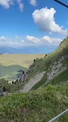 The video shows a sweeping panoramic view from a high vantage point in a mountainous region. The foreground is dominated by lush green grass and vegetation, with some of the grass blades appearing wind-blown. In the midground, a steep, grassy slope descends, revealing rocky outcrops and a scree slope. A solitary metal tower, possibly a telecommunications mast, stands on a rocky promontory. Beyond this, a dense evergreen forest carpets a large valley. Further in the distance, rolling hills with lighter green grassy tops are visible, casting shadows from scattered clouds. The background features layers of hazy blue mountains under a bright blue sky with scattered white clouds. The scene is natural and serene, with no people or animals visible. The time of day appears to be daytime, likely late morning or early afternoon, given the bright sunlight and distinct shadows.