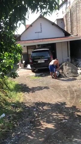 A man wearing blue shorts and no shirt is seen shoveling a pile of sand in front of a house. A dark-colored SUV is parked in the driveway. The environment appears to be a residential area with some greenery and construction materials.