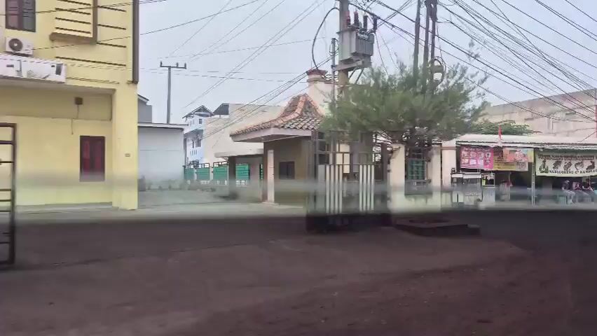 The video shows a street scene in a built-up area, likely in Indonesia, based on the script visible on signs. The time appears to be daytime, with overcast skies. The perspective is from inside a vehicle, suggesting the footage was captured while moving.

On the left, a metal gate is visible, leading into a property with a building and a tree. A power transformer is mounted on a pole. Further down the street, there are residential or commercial buildings.

The central focus of the video is a row of shops and food stalls. A prominent archway above the shops reads "GRIYA SEJAHTERA UJUNG ILIR." The shops appear to be varied, with signs indicating services like hair salons ("PANGKAS RAMBUT SRIWIJAYA"), food establishments ("MI AYAM & BAKSO", "RM. GALEKO BASALO"), and possibly a money changer ("ATM"). Several food carts or stalls are also visible.

Several vehicles are present, including a black minivan, a silver sedan, and a silver hatchback. There are also motorcycles parked along the street. People are seen walking and interacting with the stalls, including two individuals wearing dark shirts with yellow lettering and headscarves, and others in the background. The street itself is paved, and there are numerous overhead power lines. The overall impression is of a bustling, everyday commercial and residential environment.
