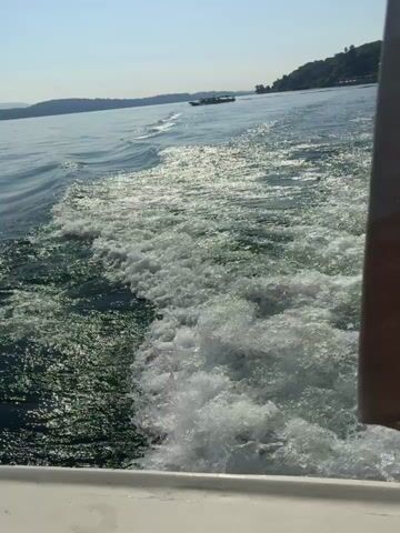 The video captures a scenic boat ride on a sunny day, likely on a lake near Binda, Italy. The camera is positioned on a moving boat, showing its white, foamy wake trailing behind on the blue-green water. In the distance, majestic mountains with some cloud cover rise from the shoreline under a clear, bright blue sky. The sun is high, casting a strong glare on the water's surface in some shots. The view alternates between the boat's wake with distant mountains and wider vistas of the tranquil lake and its mountainous surroundings. One shot is framed by the dark structure of the boat's canopy, offering a glimpse of the calm water and distant land. No people or visible text are present in the video.